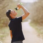 Side view of young male athlete wearing sports clothes and cap drinking water from plastic bottle on blurred background of forest during running and workout